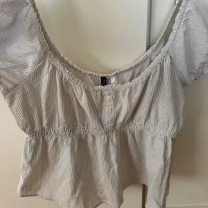 Topp från hm - Supersöt croppad blus från hm i ljusgrått och vitt med tunna ränder. Modellen har korta puffärmar, smock i midjan och knappar framtill. Perfekt för en chill och trendig look med sin elastiska halsringning och luftiga bomullsmaterial.