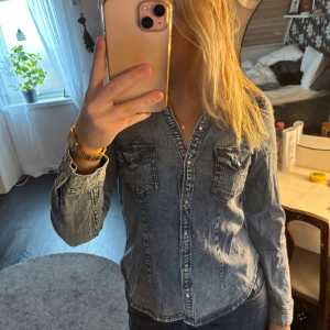 Figursydd jeansskjorta med knappar  - Säljer en snygg figursydd jeansskjorta i blått med långa ärmar och tryckknappar framtill. Skjortan har två bröstfickor med lock och markerade sömmar för en cool look. Perfekt för dig som gillar denim och vill ha en klassisk men trendig stil.