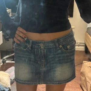 Victoria Beckham kjol - Säljer en blå jeanskjol från Victoria Beckham med klassisk femficksdesign och snygg gul broderad krona på bakfickan. Kjolen är kort och har en lätt tvättad look med slitningar för en cool vibe. Perfekt till sneakers eller boots.