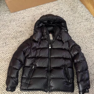 Svart Moncler Maya - Svart pufferjacka från Moncler med glansig finish och avtagbar huva. Jackan har två snedställda fickor med dragkedja, quiltad design och klassisk Moncler-serie på insidan. Perfekt för kalla dagar och har en cool streetstil. Ena fickan är tyvärr trasig , och ena ärmen är syd se bilder 