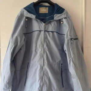 Ljusblå vindjacka från MyWear - Snygg ljusblå vindjacka från MyWear med huva och dragkedja framtill. Jackan har mörkblå detaljer, praktiska fickor med dragkedja och justerbar huva med snörning. Perfekt för blåsiga dagar och har en loose passform. Använd en gång. Storleklapp avklippt, str.46, bröst 128cm, längd 80cm.