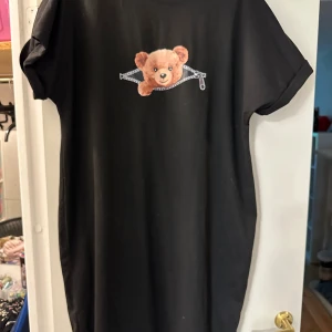 Svart t-shirtklänning med nalle från SHEIN strl: L - Svart t-shirtklänning från SHEIN i storlek L med ett gulligt tryck av ett nallehuvud som tittar ut ur ett blixtlås på bröstet. Klänningen har korta ärmar och är gjord i mjuk bomull, perfekt för en chill och avslappnad stil.