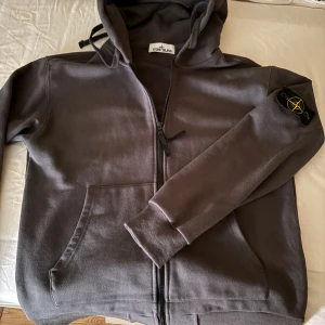 Grå hoodie från Stone Island - Säljer en grå hoodie från Stone Island med dragkedja och huva. Klassisk patch på vänster ärm och känguruficka framtill. Mjuk bomullskvalitet och ribbade muddar. Perfekt för dig som gillar streetwear och snygga detaljer. Ingår ett extra märke! 