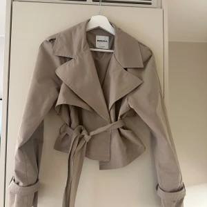 Beige kort trenchcoat från Bershka - Snygg kort trenchcoat från Bershka i beige! Populär och säljs inte längre!❤️ använd en gång 