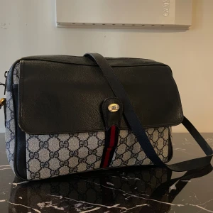 Gucci messenger i svart skinn och canvas - Snygg axelväska från Gucci med klassiskt GG-mönster i grått och svart canvas, detaljer i svart skinn och en röd/blå rand framtill. Väskan har justerbar axelrem, guldfärgad spänndetalj och dragkedja upptill. Perfekt för dig som gillar ikoniska designeraccessoarer.