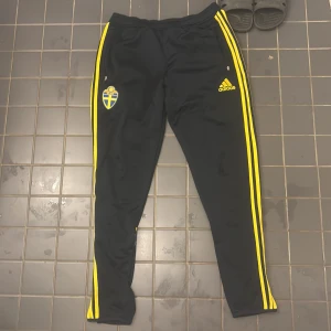 Adidas Sverige träningsbyxor svart/gul - Snygga svarta träningsbyxor från Adidas med gula ränder längs sidorna och broderad Sverige-logga (SVFF) på låret. Byxorna har dragkedjor vid fickorna och detaljer i blått och gult på baksidan av benen. Perfekta för fotbollsträning eller chill.