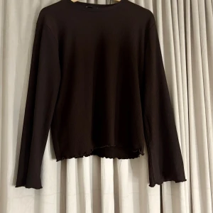 Brun långärmad topp med vågig kant - Mörkbrun långärmad topp i ribbad bomullsmix med vågiga kanter vid ärmslut och nederkant. Enkel och stilren modell med rund halsringning och mjukt, stretchigt material som sitter snyggt på kroppen.