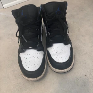 Nike Air Jordan 1 Mid svart/blå/vit - Nike Air Jordan 1 Mid i svart, blått och vitt skinn. Klassisk siluett med blå swoosh och detaljer, svart snörning och perforerad tå. Ikonisk Jordan-logga på sidan och tungan. Perfekta sneakers för dig som gillar streetstyle.