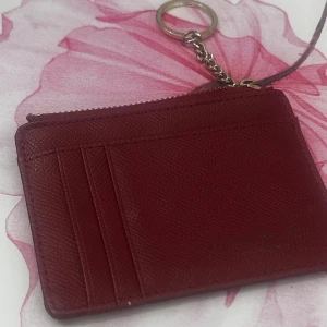 Vinröd plånbok med nyckelring - Snygg vinröd plånbok i skinn med plats för kort och mynt. Praktisk dragkedja upptill och en kedja med nyckelring så du enkelt kan fästa den i väskan eller nyckelknippan. Perfekt storlek för att ha med sig överallt.