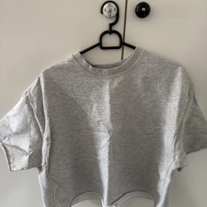 Grå croppad topp från Zara - Säljer en ljusgrå croppad topp från Zara i mjuk bomull. T-shirten har rund halsringning, korta ärmar och en loose passform. Perfekt att matcha med höga jeans eller shorts för en chill look.