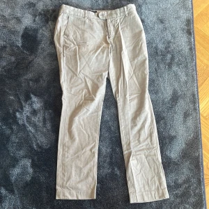Beige chinos från Brukslid, stl 31 - Snygga beige chinos från Brukslid i storlek 31. Klassisk rak passform med bälteshällor, sidfickor och bakfickor med knappar. Tillverkade i mjuk bomull, perfekta för en clean och stilren look.