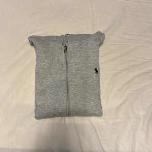 Grå hoodie från Polo Ralph Lauren - Grå hoodie från Polo Ralph Lauren med dragkedja framtill och klassisk svart logga broderad på bröstet. Tröjan har huva med snörning, två fickor och långa ärmar. Perfekt för en chill och stilren look.