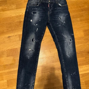 Mörkblå distressed jeans från Dsquared2 - Säljer ett par mörkblå jeans från Dsquared2 med slitna detaljer, hål och färgstänk för en riktigt edgy look. Klassisk femficksmodell med raka ben och snygg tvätt. Jeansen är i bomull med lite stretch för skön passform.