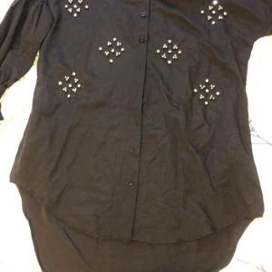 Svart skjorta med pärldetaljer - Svart långärmad skjorta med snygga pärldetaljer framtill. Skjortan har knappar hela vägen och en lite längre, rundad nederkant. Ärmarna har knytband vid handlederna för extra stilpoäng. Perfekt för dig som vill sticka ut lite extra.