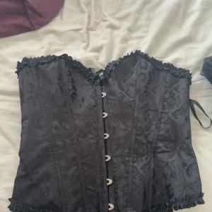 Svart korsett med snörning och mönster - Säljer en svart korsett med snyggt tonat blommigt mönster och dekorativa volanger längs kanterna. Framsidan har metallknäppning och baksidan har snörning för att justera passformen. Materialet är glansigt och känns lyxigt, perfekt för att lyfta din outfit. Helt ny aldrig använd