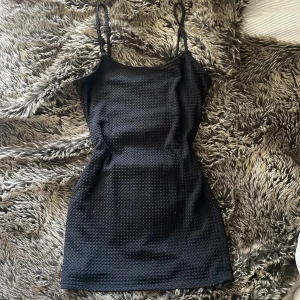 Svart kort klänning från H&M Divided - Säljer en svart kort klänning från H&M Divided i storlek S. Klänningen har smala axelband och ärmlös design, med ett coolt texturerat tyg och slät insida. Sitter snyggt och har en slim passform som framhäver figuren.