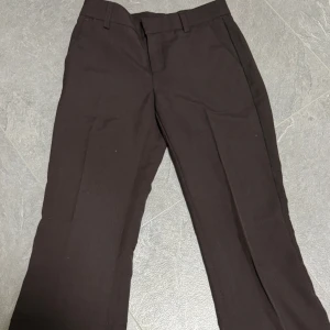 Bruna kostymbyxor från Gina Tricot - Snygga bruna kostymbyxor från Gina Tricot i storlek 32. Byxorna har klassisk rak passform, hällor för bälte och pressveck framtill. Perfekta för en clean och stilren look. Materialet känns mjukt och följsamt.