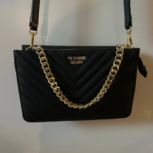 Svart axelväska från Victoria's Secret - Snygg svart axelväska från Victoria's Secret med quiltad yta och guldfärgad kedjedetalj. Väskan har justerbar axelrem och dragkedja upptill. Insidan är fodrad med svart randigt tyg och har logga invändigt. Perfekt för att lyfta din outfit. Helt i nyskick 