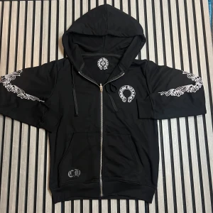 Svart hoodie Chrome Hearts x Comme des Garçons - Svart hoodie med dragkedja och huva från Chrome Hearts x Comme des Garçons. Snygga vita tryck på bröstet, ryggen och längs ärmarna med ikoniska loggor och detaljer. Tröjan har snörning vid huvan och ribbade muddar. Perfekt för dig som gillar streetwear med edge.