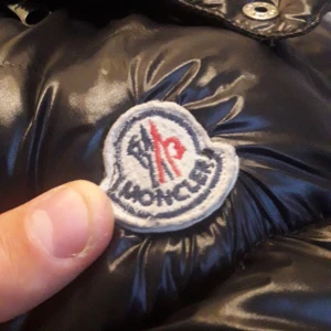 Mer bilder på moncler jackan! - Säljer en svart, glansig dunjacka från Moncler med huva och dragkedja framtill. Jackan har ett quiltat mönster och klassisk Moncler-logga på bröstet. Fylld med vitt gåsdun för extra värme. Perfekt för kalla dagar och riktigt snygg streetstil.
