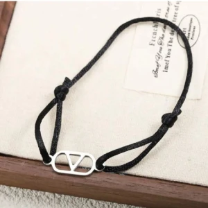 Svart armband med silverfärgad detalj - Stilrent armband med svart justerbart band och en silverfärgad metallbricka i form av en oval med V-liknande design. Perfekt för dig som gillar minimalistiska accessoarer med en modern touch.