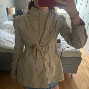 Beige trench coat med huva och snörning - Snygg beige parkajacka med huva och hög krage. Jackan har dragkedja framtill, knappar och snörning i midjan för en schysst passform. 