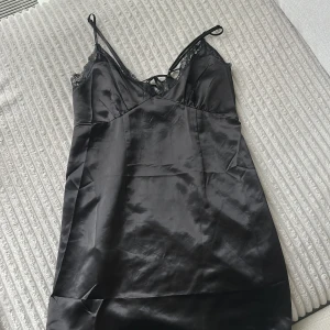 Svart satinklänning - Svart kort klänning  i satin med tunna axelband, spetsdetaljer vid urringningen och snörning i bak. Klänningen har glansigt tyg