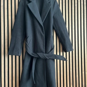 Svart kappa från Vero Moda med bälte - Stilren svart kappa från Vero Moda med klassisk krage och knytbälte i midjan. Långärmad modell i mjukt material som ger en snygg siluett. Perfekt för dig som vill ha en enkel men trendig look.Lite nopprig men annars i bra skick. Snabb leverans!!