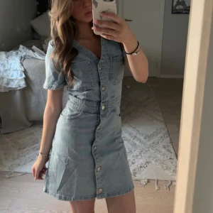 Ljusblå jeansklänning med knappar - Säljer en ljusblå jeansklänning med kort ärm och v-ringning. Klänningen har knappar hela vägen framtill och två bröstfickor. Snygg passform som markerar midjan och ger en avslappnad men trendig look. Använt 2 gånger så skicket är som ny!