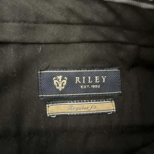 Svarta kostymbyxor från Riley - Säljer ett par svarta kostymbyxor från Riley i regular fit. Byxorna har klassisk rak passform och är tillverkade i ett mjukt material som känns bekvämt. Perfekta för dig som gillar stilrena och tidlösa plagg. Stängs med dragkedja och knapp.