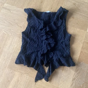 Svart volangblus från Gina Tricot - Säljer en svart blus från Gina Tricot med coolt skrynkligt tyg och volangdetaljer längs halsringningen och nederkanten. Blusen är ärmlös och har en snygg omlottkänsla framtill. Perfekt för dig som gillar unika toppar med extra detaljer. Pris kan diskuteras 