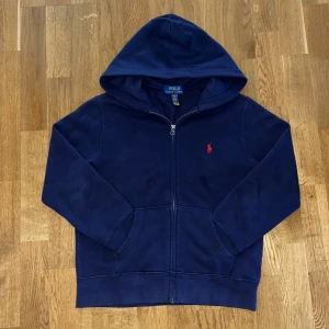 Marinblå hoodie från Polo Ralph Lauren - Snygg marinblå hoodie från Polo Ralph Lauren med röd logobrodyr på bröstet. Tröjan har huva, dragkedja framtill och två fickor. Tillverkad i mjuk bomullsmix med fleecefoder på insidan för extra komfort. Perfekt för chill dagar och skön stil.
