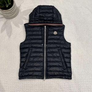 Moncler gilet  - Svart dunväst från Moncler med huva och klassisk logga på bröstet. Västen har quiltad design, dragkedja framtill och två snedställda fickor. Snygg detalj med röd, vit och blå rand längs huvkanten. Perfekt för lager-på-lager-stil.