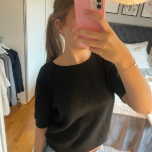 Svart stickad topp från H&M - En svart stickad topp från H&M med korta ärmar och rund halsringning. Modellen är croppad och har en mjuk, lite luddig känsla. Perfekt att styla off-shoulder för en chill vibe. Passar till jeans eller kjol för en enkel men snygg look.