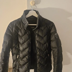 Rockandblue svart vinterjacka  - Snygg svart pufferjacka från Rockandblue med quiltat mönster och hög krage. Jackan har dragkedja framtill, två fickor med dragkedja och en patch på ena ärmen. Perfekt för kyliga dagar och riktigt stilren look.