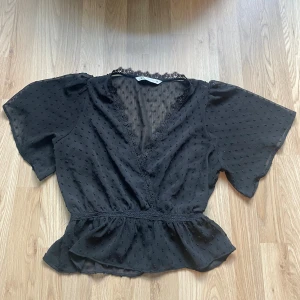 Svart blus med spets från Zara - Supersöt svart blus från Zara med v-ringning och spetsdetaljer längs halsen. Blusen har korta, vida ärmar och ett diskret prickigt mönster i tyget. Blusen har ett litet hål (se bild) men ingenting som syns när man har den på sig! 