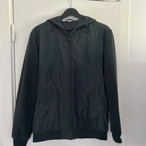 Grön cardigan från Massimo Dutti - Grön cardigan från Massimo Dutti med huva och snörning. Jackan har dragkedja framtill, två sidofickor och ribbade muddar vid ärmslut och nederkant. Materialet är en mix av polyester och bomull, vilket gör den lätt och bekväm. Perfekt för dig som gillar stilren och enkel design.