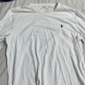 Vit långärmad t-shirt Polo Ralph Lauren - Vit långärmad t-shirt från Polo Ralph Lauren i mjuk bomull. Klassisk rund hals och diskret blå logga broderad på bröstet. Perfekt för dig som gillar stilrena och enkla plagg med en touch av lyx.