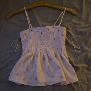 Ljus blommig topp   - Supersöt ljus linnetopp från Gina med smockad överdel och tunna axelband. Toppen har små blommor i rosa och lila, och ett lätt rynkat fall nedtill. 