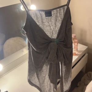 Grå linnetopp med knyt från Gina Tricot - Säljer en grå linnetopp från Gina Tricot med smala axelband och snygg knytning framtill. Toppen har en v-ringad design och är tillverkad i mjuk bomull, perfekt för en chill och stilren look.
