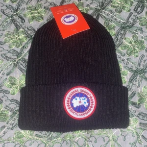 Canada Goose mössa - Svart ribbstickad mössa från Canada Goose med klassisk uppvikt kant och det ikoniska runda Arctic Program-märket framtill. Tillverkad i mjukt och värmande material, perfekt för kyliga dagar. Snygg och enkel design som funkar till allt.