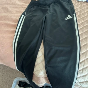 Svart Adidas sett - Svart Adidas zip-tröja med klassiska vita ränder längs ärmarna och broderad logga på bröstet. Tröjan har dragkedja framtill och två fickor med dragkedja. Tillverkad i mjuk polyester, perfekt för träning eller chill. Svarta adidas byxor, säljs som ett sett eller sepparat.