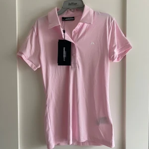 Ljusrosa polo t-shirt J.Lindeberg  - Snygg ljusrosa polo t-shirt från J.Lindeberg i stretchigt funktionsmaterial. Klassisk krage, knappar framtill och diskret logga på bröstet. Perfekt för dig som gillar sportig och stilren look. Materialet är snabbtorkande och andas. 