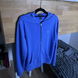 Blå hoodie från Polo Ralph Lauren XL - Blå hoodie från Polo Ralph Lauren i storlek XL. Tröjan har dragkedja framtill, huva och två fickor. Klassisk gul broderad logga på bröstet. Perfekt för chill dagar och snygg till jeans eller joggers.