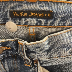 Blå jeans från Nudie Jeans Co - Säljer ett par klassiska blå jeans från Nudie Jeans Co med snyggt tvättad finish och kontrastsömmar. Jeansen har raka ben, normal passform och ikoniska bakfickor med broderad våg. Perfekta för en avslappnad streetstil.