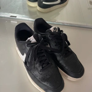 Svarta Nike Court sneakers - Svarta Nike Court sneakers med vit sula och vit swoosh på sidorna. Skorna har klassisk snörning, rund tå och är tillverkade i skinn med meshfoder för extra komfort. Perfekta för dig som gillar en stilren och sportig look.