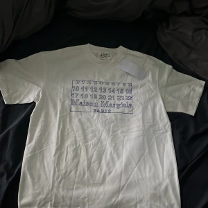 Maison Margiela vit t-shirt med print - Sprillans ny Vit t-shirt från Maison Margiela med klassiskt tryck framtill med siffror och logga i blått. Modellen har rund hals och korta ärmar. Materialet är mjuk bomull och passformen är normal. Perfekt för dig som gillar stilrena designer med en twist.