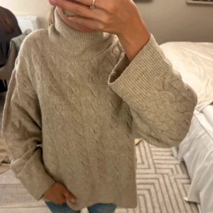 Beige stickad polotröja med kabelmönster - Mysig beige stickad polotröja med klassiskt kabelstickat mönster. Tröjan har en hög polokrage, breda muddar och en loose passform som ger en avslappnad vibe. Perfekt för kyliga dagar och enkel att styla med jeans eller kjol.