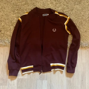 Vinröd Fred Perry kofta med gula ränder - Säljer en vinröd kofta från Fred Perry med dragkedja och vita samt gula ränder på axlar och muddar. Klassisk logga broderad på bröstet. Perfekt för dig som gillar sportig och retro stil.