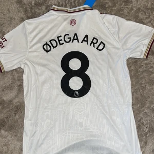Arsenal Ødegaard 8 Adidas fotbollströja M - Snygg Arsenal fotbollströja från Adidas med Ødegaard och nummer 8 på ryggen. Tröjan är vit med vinröda och guldiga detaljer, korta ärmar och klassisk krage. Broderade loggor och diskret mönster i tyget. Materialet är lätt och andas, perfekt för match eller träning.